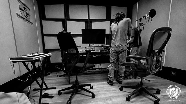 Black-and-white photo of Luigui Bleand, seen from behind, at the desk of a recording room at Universal Music Studios in Milan, beside a microphone; his crest in the corner