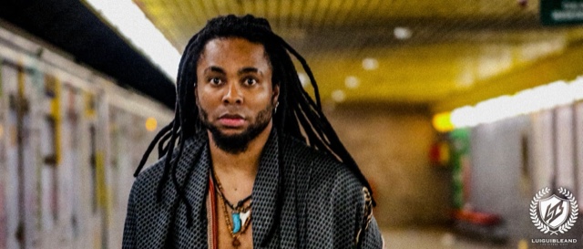 Colour photograph of Luigui Bleand walking toward the camera through an underground corridor, in a dark patterned jacket and necklaces, the corridor behind him out of focus
